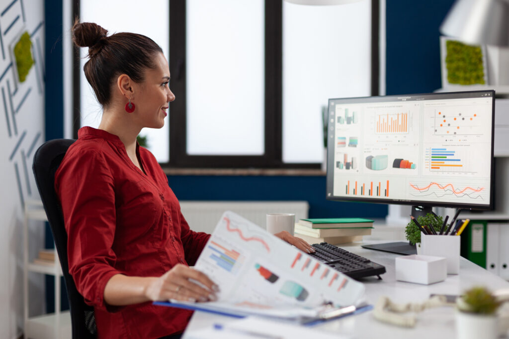 businesswoman-with-clipboard-sitting-desk-startup-business-office-smiling-employee-red-shirt-comparing-charts-successful-entrepreneur-looking-desktop-computer-analyze-business-data