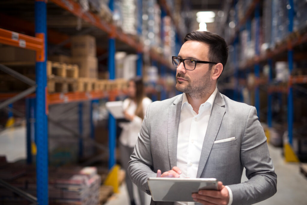 portrait-successful-businessman-manager-ceo-holding-tablet-walking-through-warehouse-storage-area-looking-towards-shelves